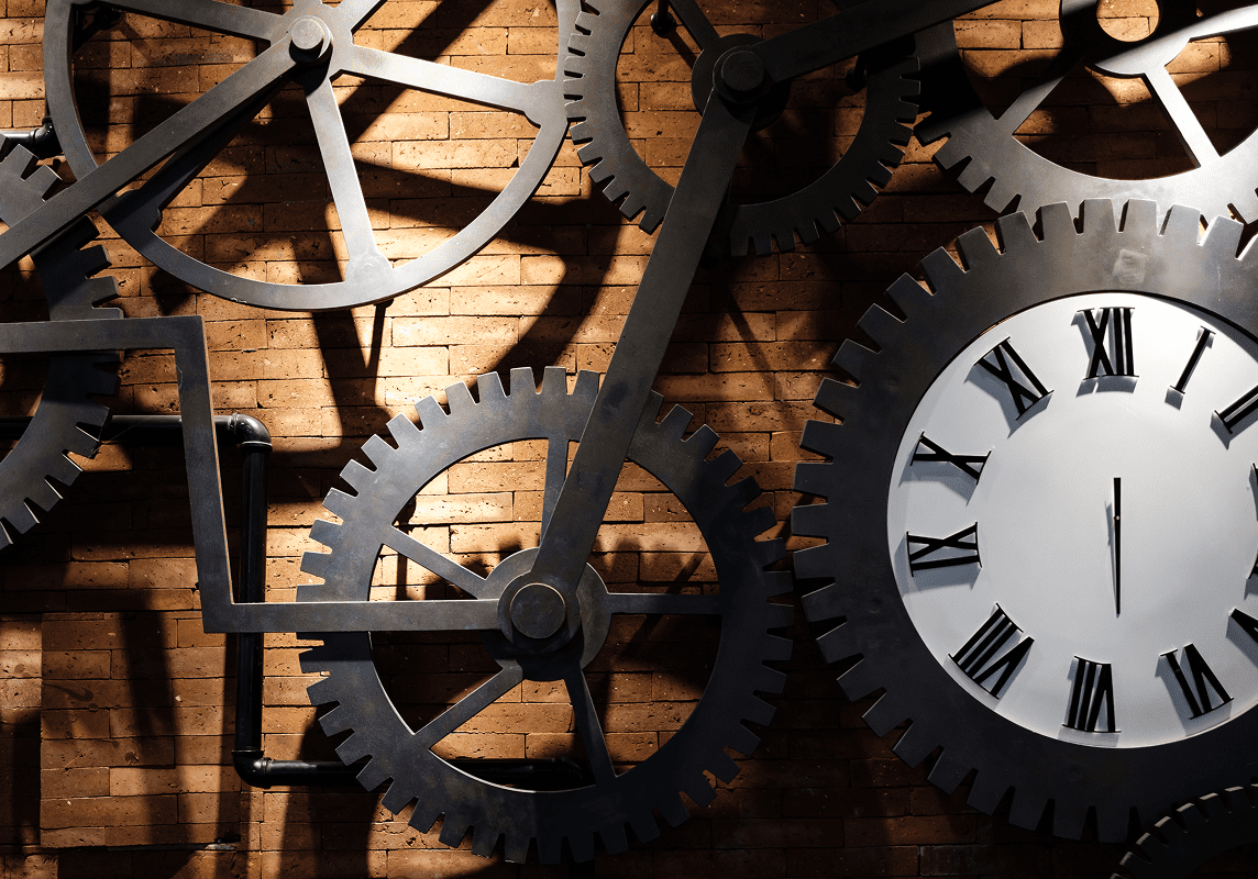 Large metal gears and a clock with Roman numerals are mounted on a brick wall, illuminated by overhead light.